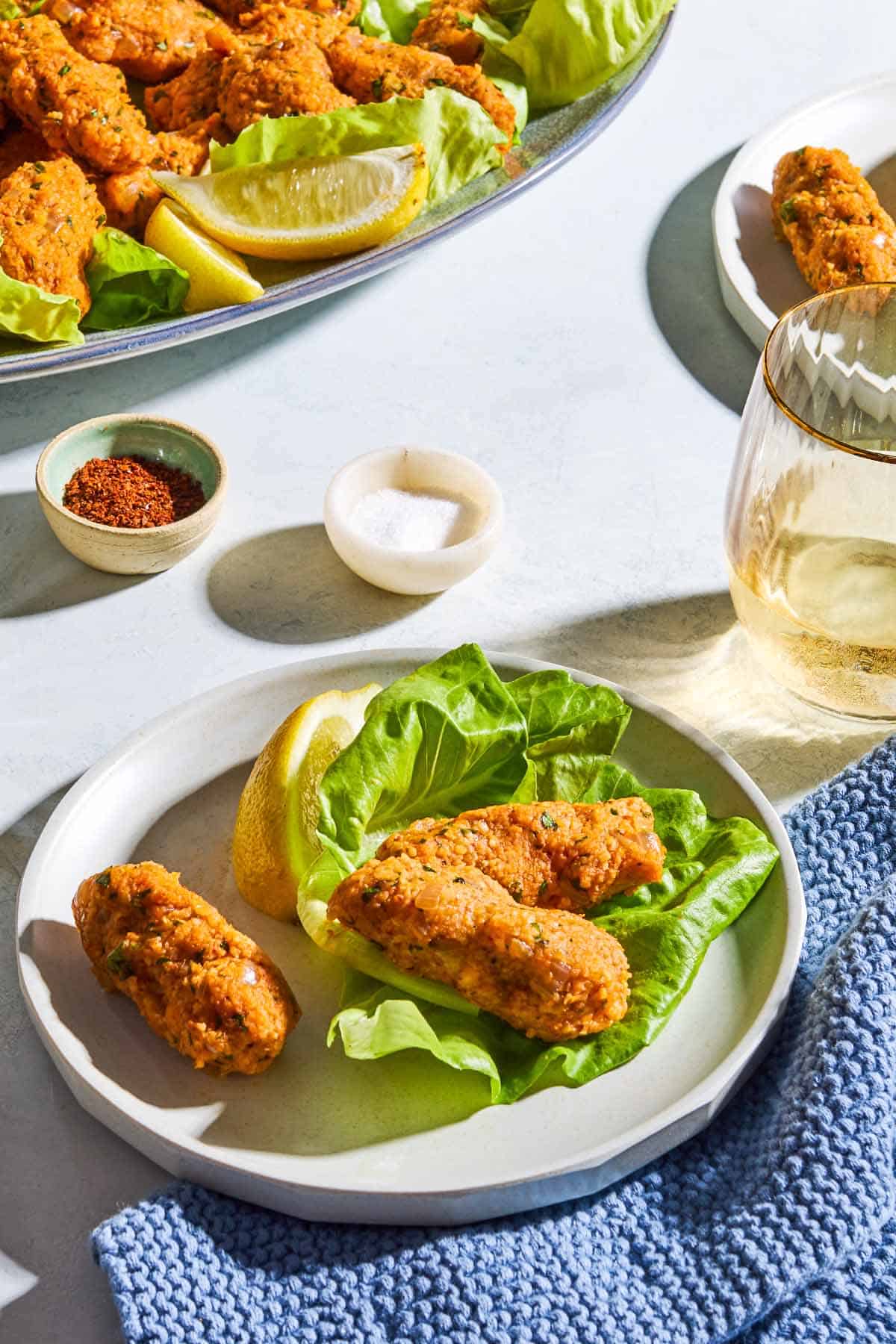 3 turkish lentil meatballs called Mercimek Köftesi on a plate with a lemon wedge, 2 of them are in a lettuce boat. In the background are bowls of salt and aleppo pepper, a glass of white wine, and the rest of the patties on serving platter with more lettuce and lemon wedges.