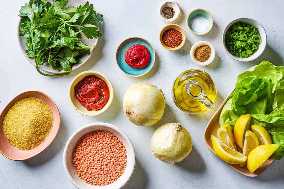 Ingredients for turkish lentil meatballs called Mercimek Köftesi including red lentils, olive oil, onions, tomato paste, turkish red pepper paste, fine bulgur, green onions, parsley, cumin, aleppo pepper, salt, black pepper, lettuce cups, and lemon wedges.