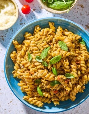 An overhead photo of pesto alla trapanese pasta in a bowl surrounded by 3 cherry tomatoes and bowls of pecorino romano cheese, and basil and mint.