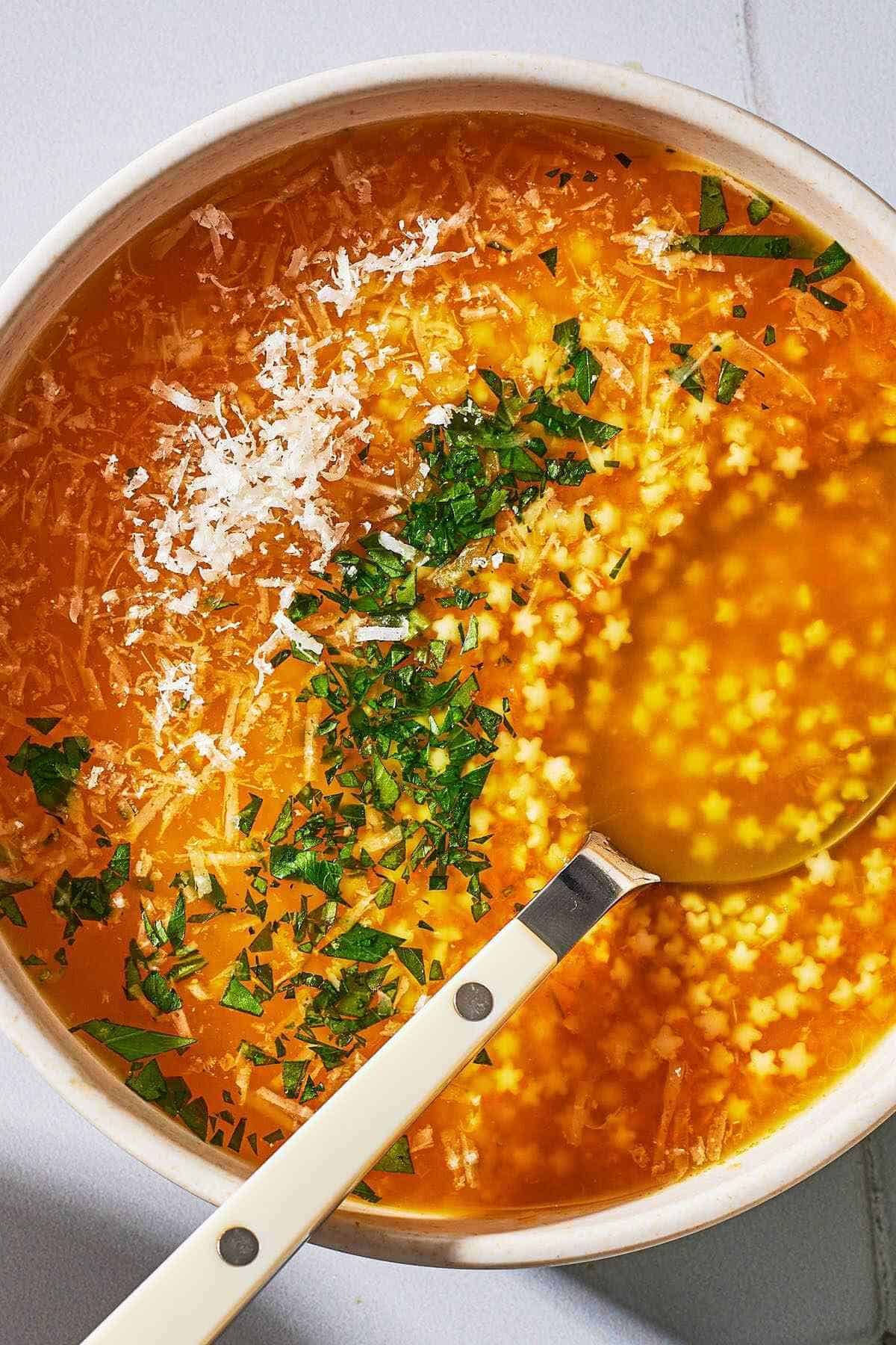 an overhead photo of a serving of pastina soup garnished with parsley and parmesan cheese in a bowl with a spoon.