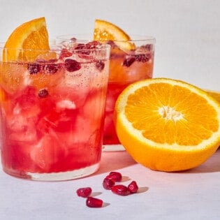 A close up of 2 pomegranate fizz mocktails garnished with pomegranate seeds and orange wedges next to more pomegranate seeds and 2 orange halves.