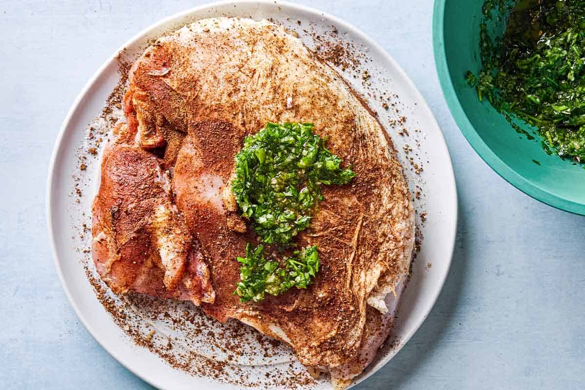 An overhead photo of an uncooked, bone-in turkey breast seasoned with spices and topped with a garlic parsley mixture on a plate. Next to this is a bowl with the rest of the garlic and parsley mixture.