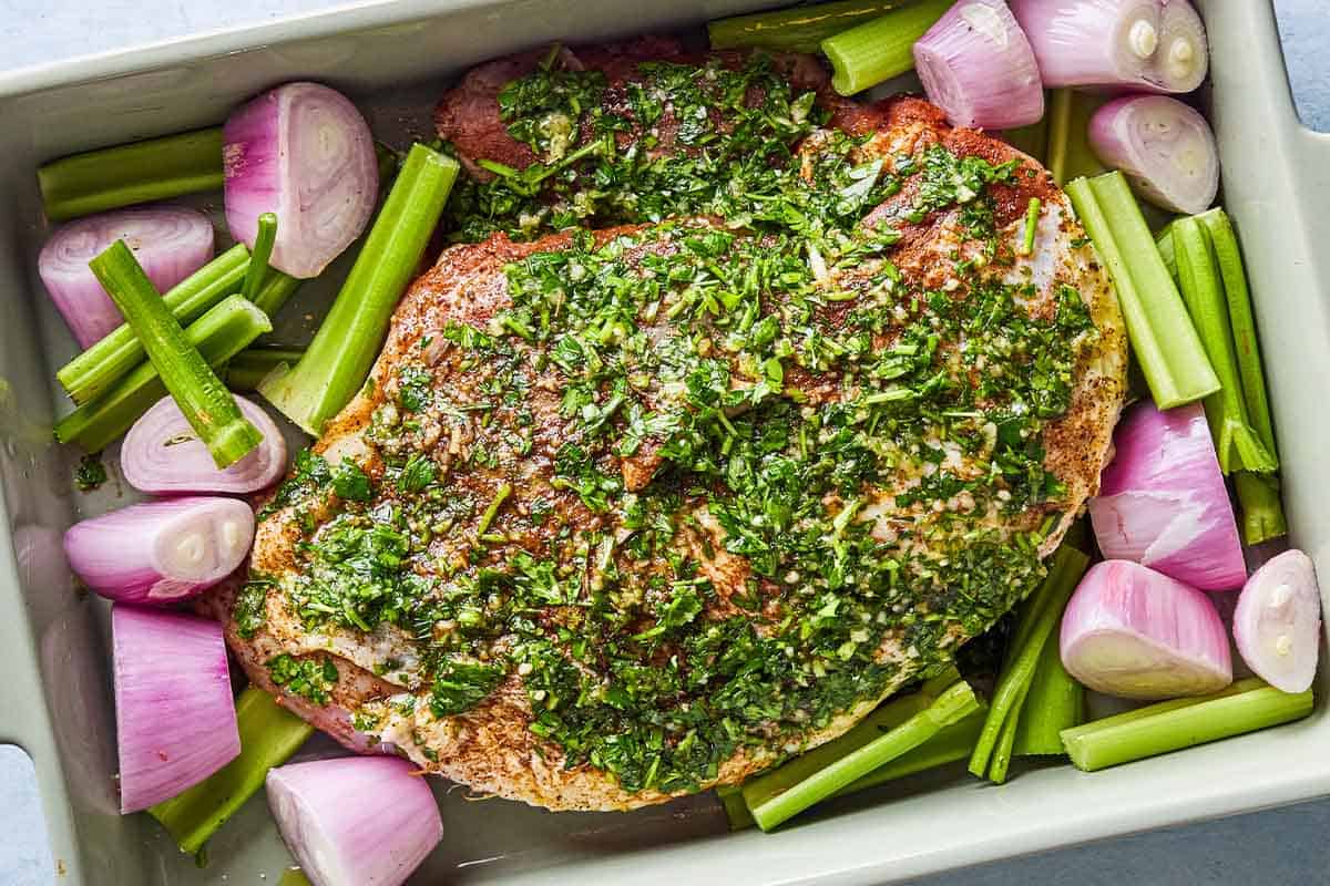 An overhead photo of a seasoned, uncooked turkey breast with shallots and celery in a baking dish.