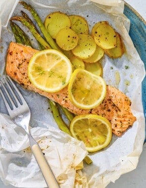 Salmon en papillote topped with lemon wheels on a bed of asparagus and potatoes on a plate with a fork.