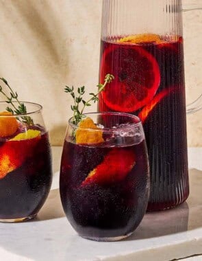 A close up of a pitcher non alcoholic sangria next two glasses of the sangria garnished with sprigs of fresh thyme and orange and lemon peels on a marble serving tray. Behind this is a bowl of oranges.