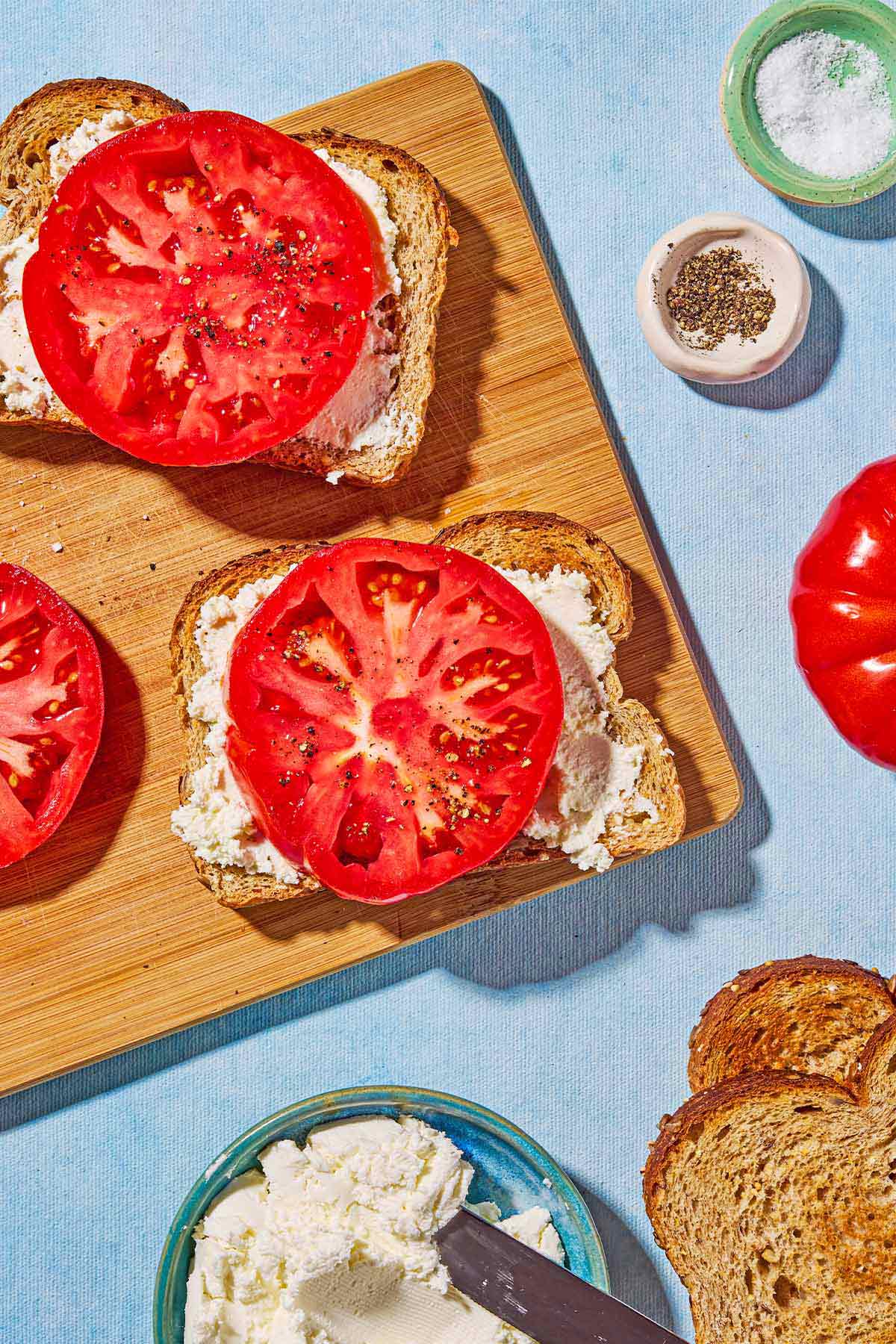 An overhead photo of two slices of toast topped with labneh, a tomato and salt and pepper next to another slice of tomato on a cutting board. Next to the cutting board is an heirloom tomato, bowls of salt and pepper, 2 pieces of toast and a bowl of whipped labneh with a knife.