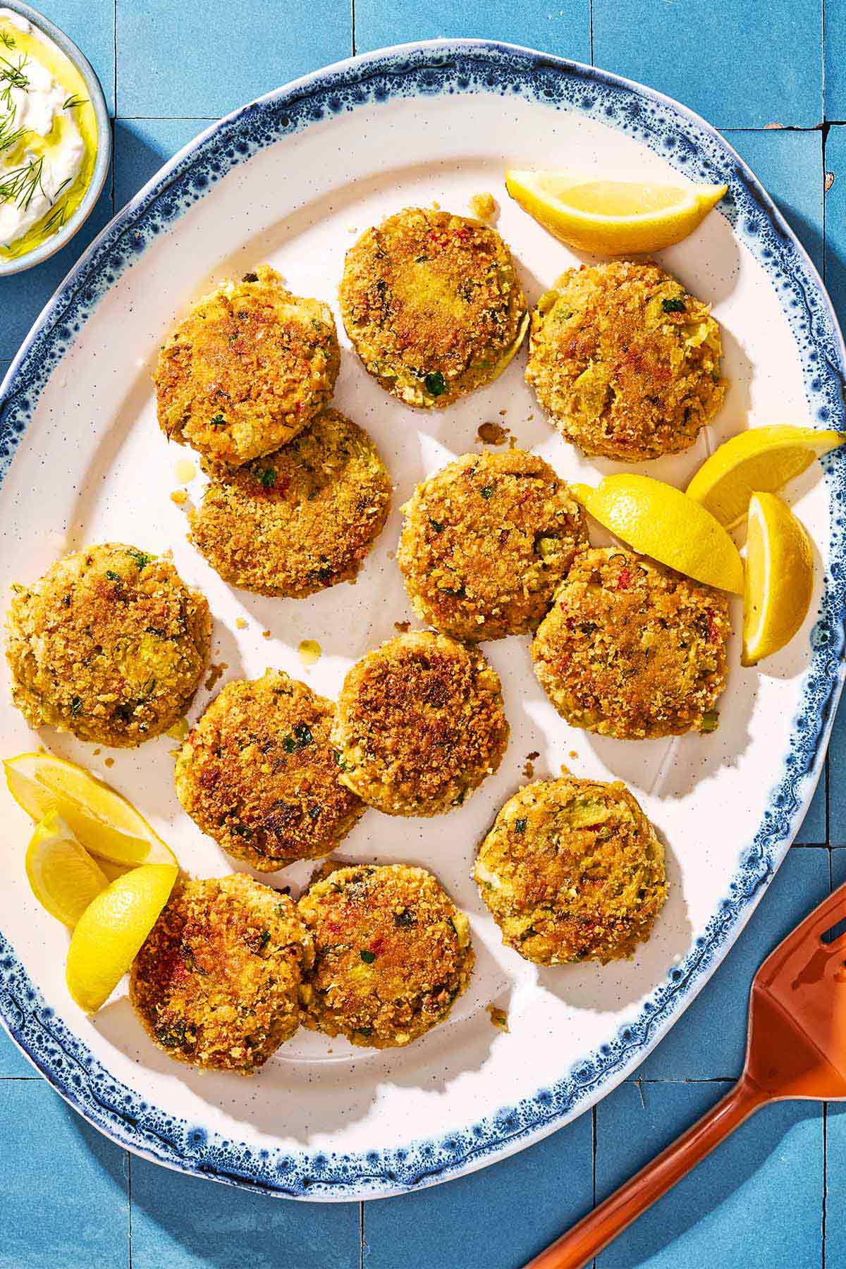 An overhead photo of vegetarian crab cakes on a serving platter with lemon wedges. Next to this is a bowl of tzatziki and a spatula.