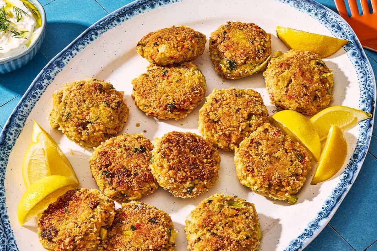 A closeup of vegetarian crab cakes on a serving platter with lemon wedges. Next to this is a bowl of tzatziki and a spatula.