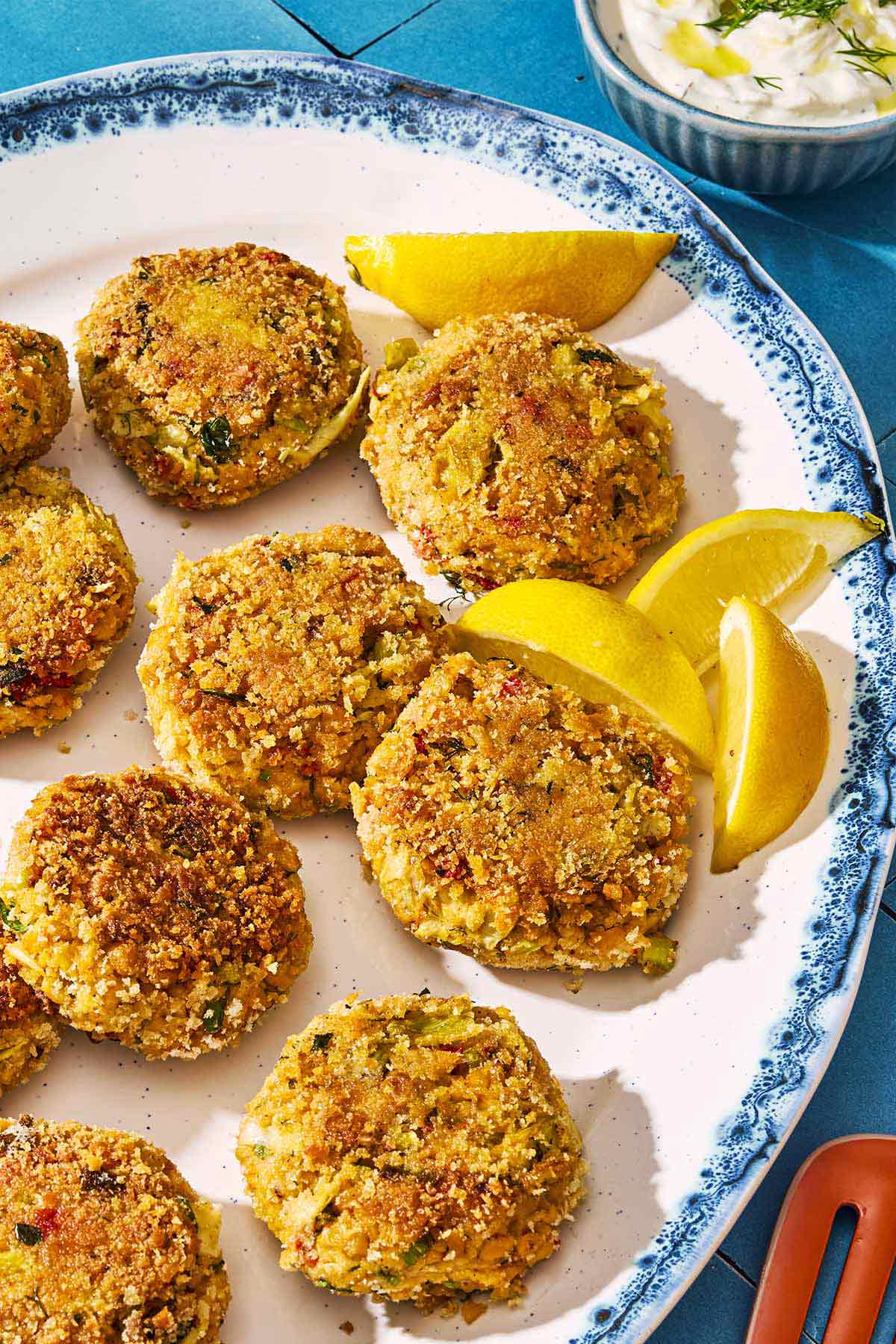 A closeup of vegetarian crab cakes on a serving platter with lemon wedges. Next to this is a bowl of tzatziki.