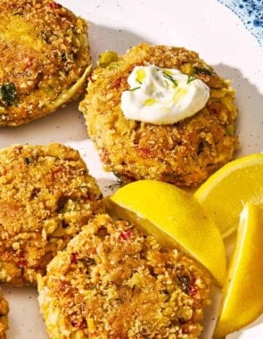 A closeup of vegetarian crab cakes on a serving platter with lemon wedges. One cake as a small dollop of tzatziki on it.