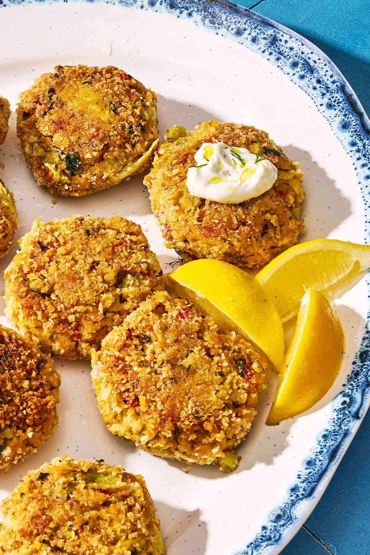 A closeup of vegetarian crab cakes on a serving platter with lemon wedges. One of the cakes has a dollop of tzatziki on it.