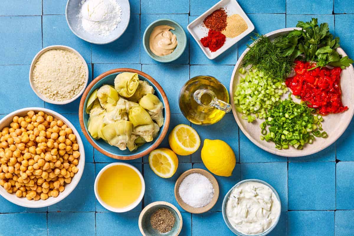 Ingredients for vegetarian crab cakes including chickpeas, artichoke hearts, breadcrumbs, scallions, celery, roasted red peppers, cornstarch, parsley, dill, dijon mustard, lemon, garlic, aleppo pepper, kosher salt, black pepper, and tzatziki sauce.