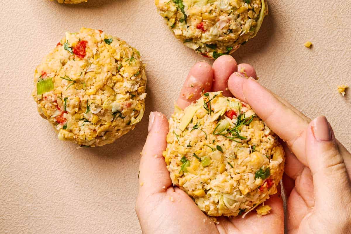 An overhead photo of a vegetarian crab cake being formed with 2 hands above 2 vegetarian crab cakes that are already formed.