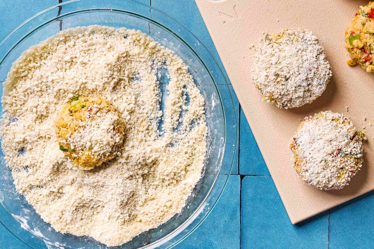 An overhead photo of an cooked crab cake being coated on a plate. Next to this is 2 coated crab cakes and one uncoated crab cake on a cutting board.