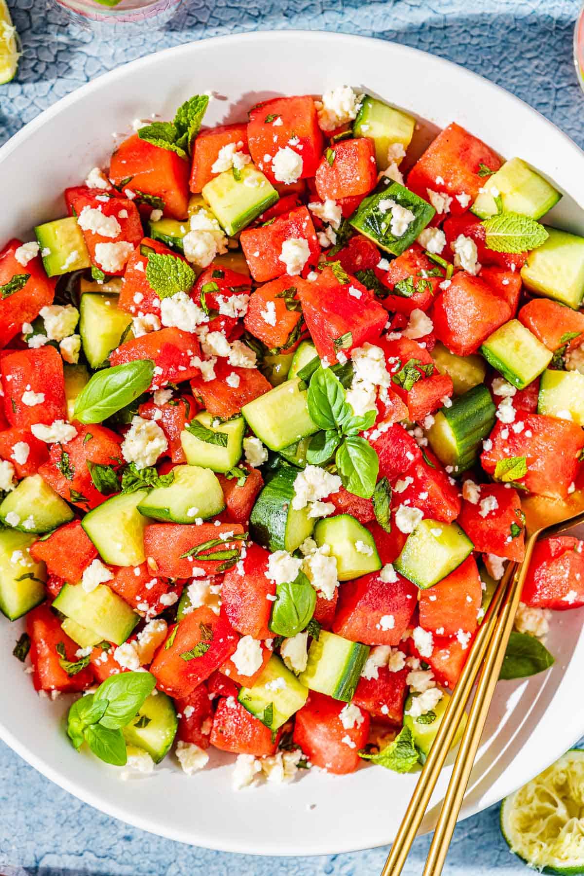 close up of a bowl of watermelon salad in a bowl with gold serving utensils.