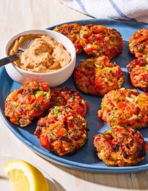 A close up of tomato fritters and a bowl of sun dried tomato yogurt dip with a spoon on a serving platter. Next to this is a plate with lemon wedges and a towel.