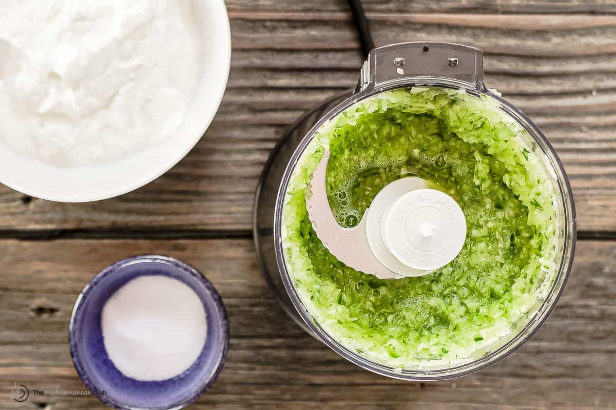 grated cucumbers in a food processor with a side of yogurt and kosher salt