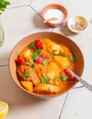 A bowl of vegetable stew with a spoon next to small bowls of salt and pepper.