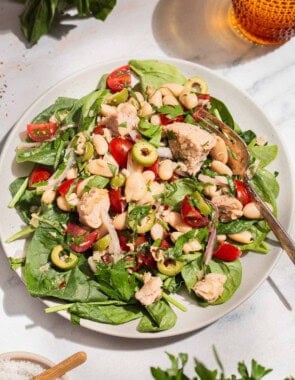 A close up photo of a serving tuna white bean salad on a plate with a fork. Next to this is a glass of water, sprigs of basil and parsley, and a bowl of kosher salt.