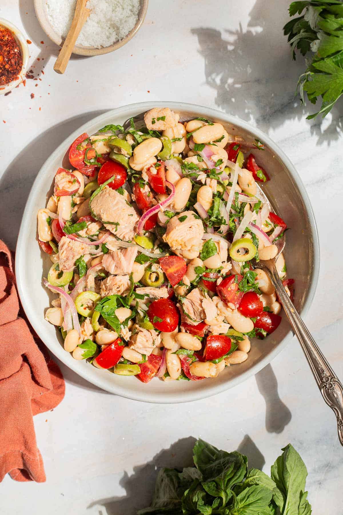 An overhead photo of tuna white bean salad in a serving bowl with a spoon. Next to this is a cloth napkin, sprigs of basil and parsley, and bowls of aleppo pepper and kosher salt.