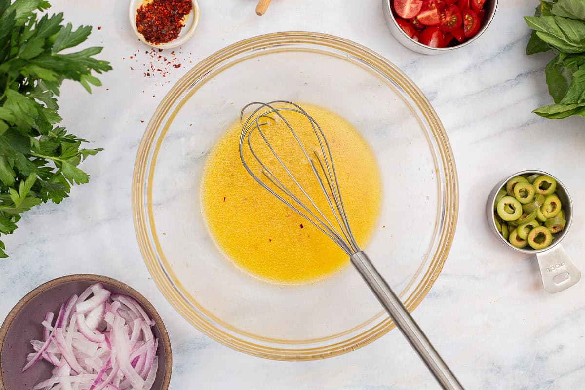 The dressing for the tuna white bean salad in a mixing bowl with a whisk. Next to this is a bowl of red onions, measuring cups with cherry tomatoes and green olives, a bowl of kosher aleppo pepper, and sprigs of basil and parsley.