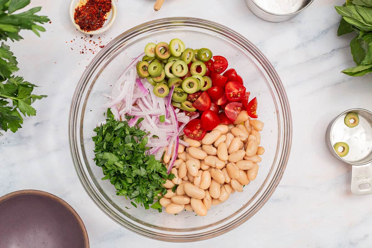 An overhead photo of the ingredients for a tuna white bean salad in a mixing bowl just before being mixed together. Next to this is an empty bowl, a bowl of aleppo pepper, sprigs of basil and parsley, and empty measuring cups.